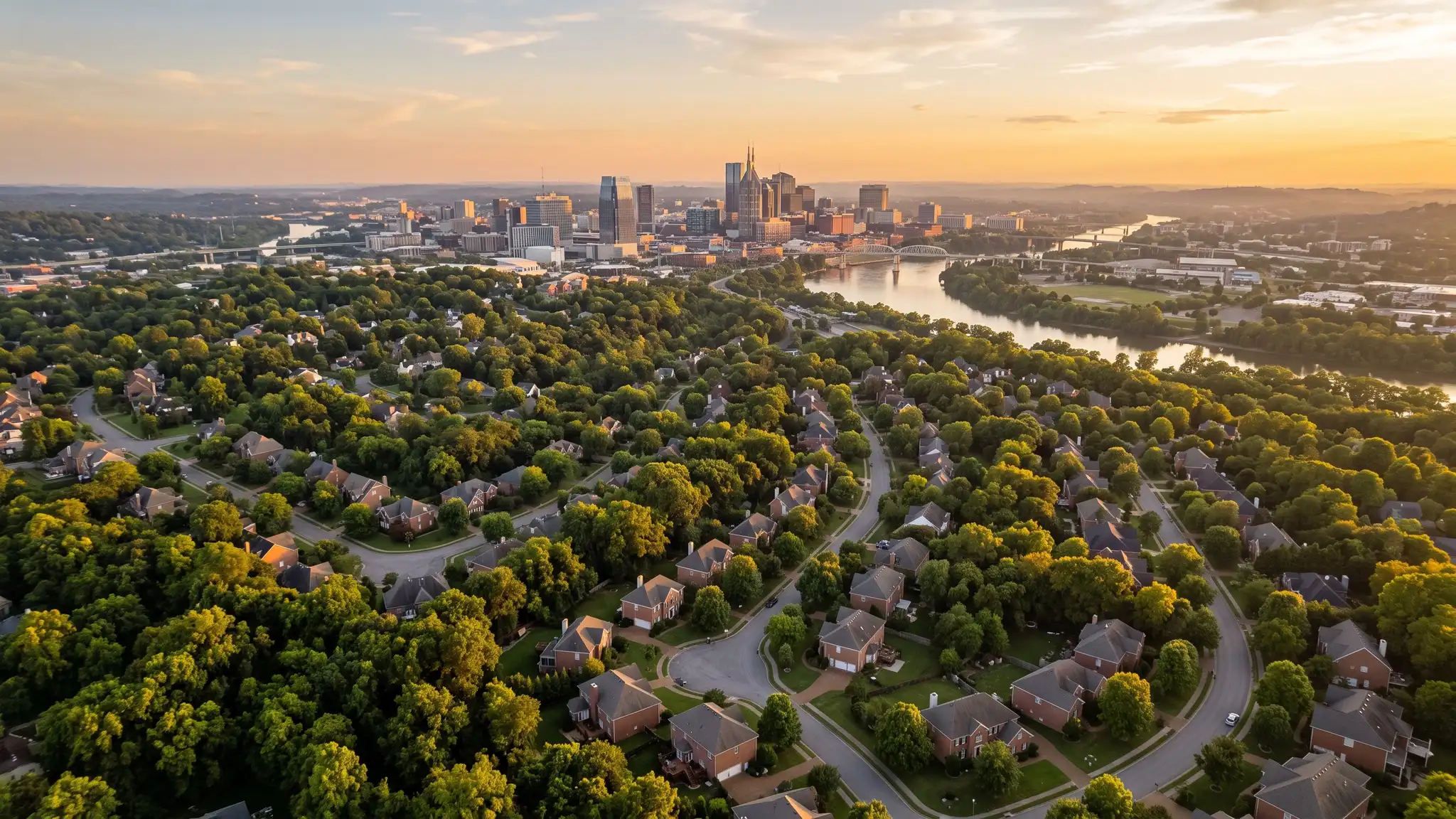 Aerial view of the Nashville skyline at golden hour above tree-covered neighborhoods with brick homes and the Cumberland River in the distance